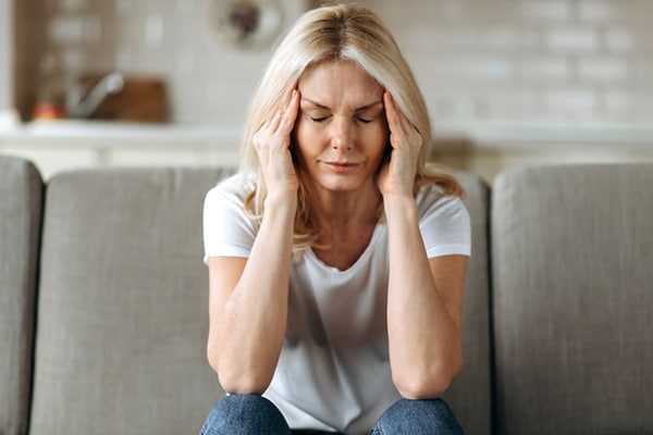 Middle aged blonde woman sits on couch at living room holding her head with her hands, feels unhappy because of headache, personal troubles, illness or bad news, she need psychological or medical support